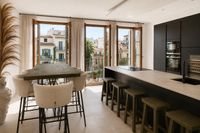 Open-plan kitchen and dining area in a Santa Catalina townhouse in Palma with oak-toned dining table, modern black kitchen, soft neutral textures and Mediterranean city views through large windows.